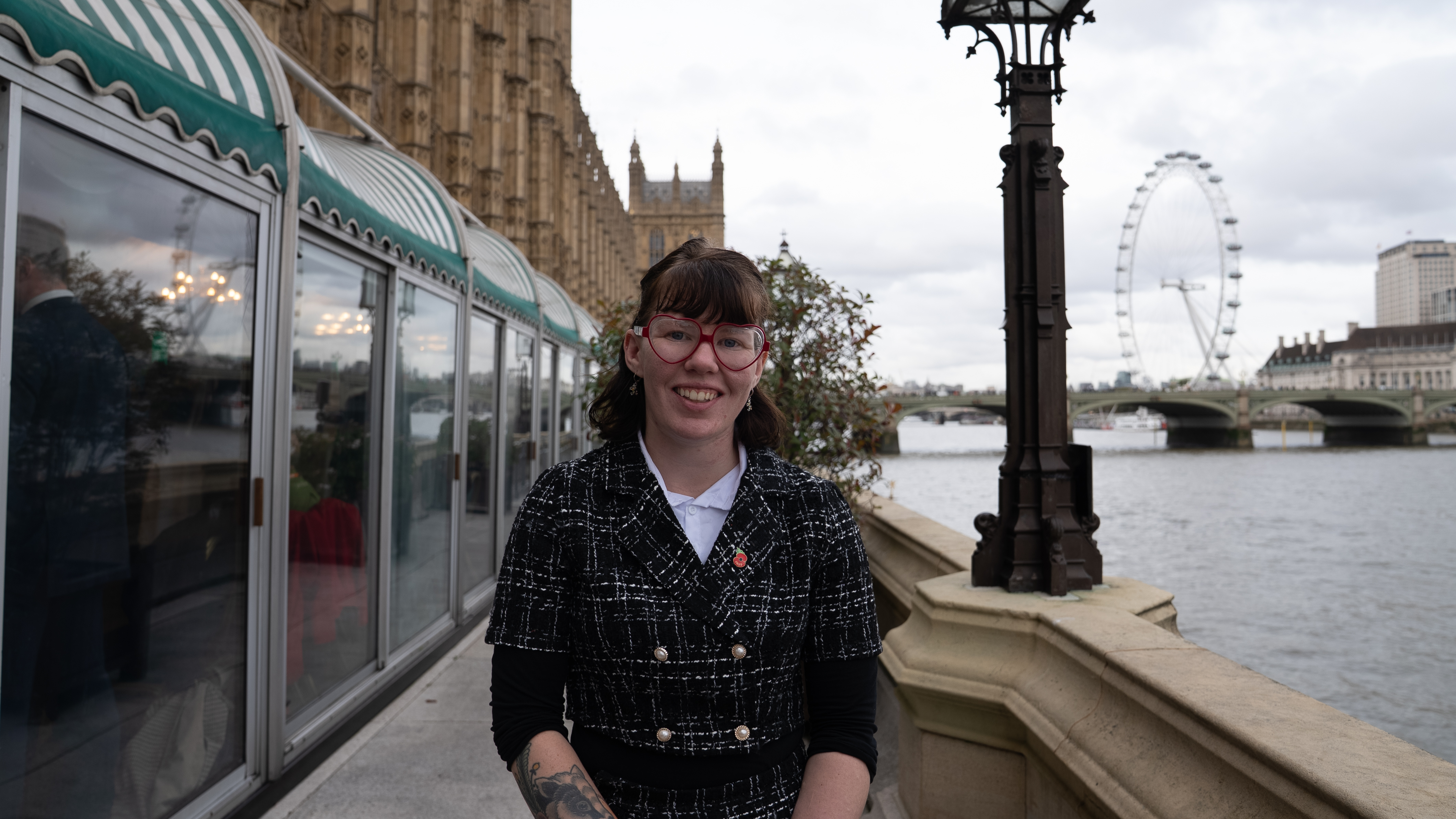 Kerrie wears red glasses, a white shirt, and a black coat, standing in front of the Houses of Parliament