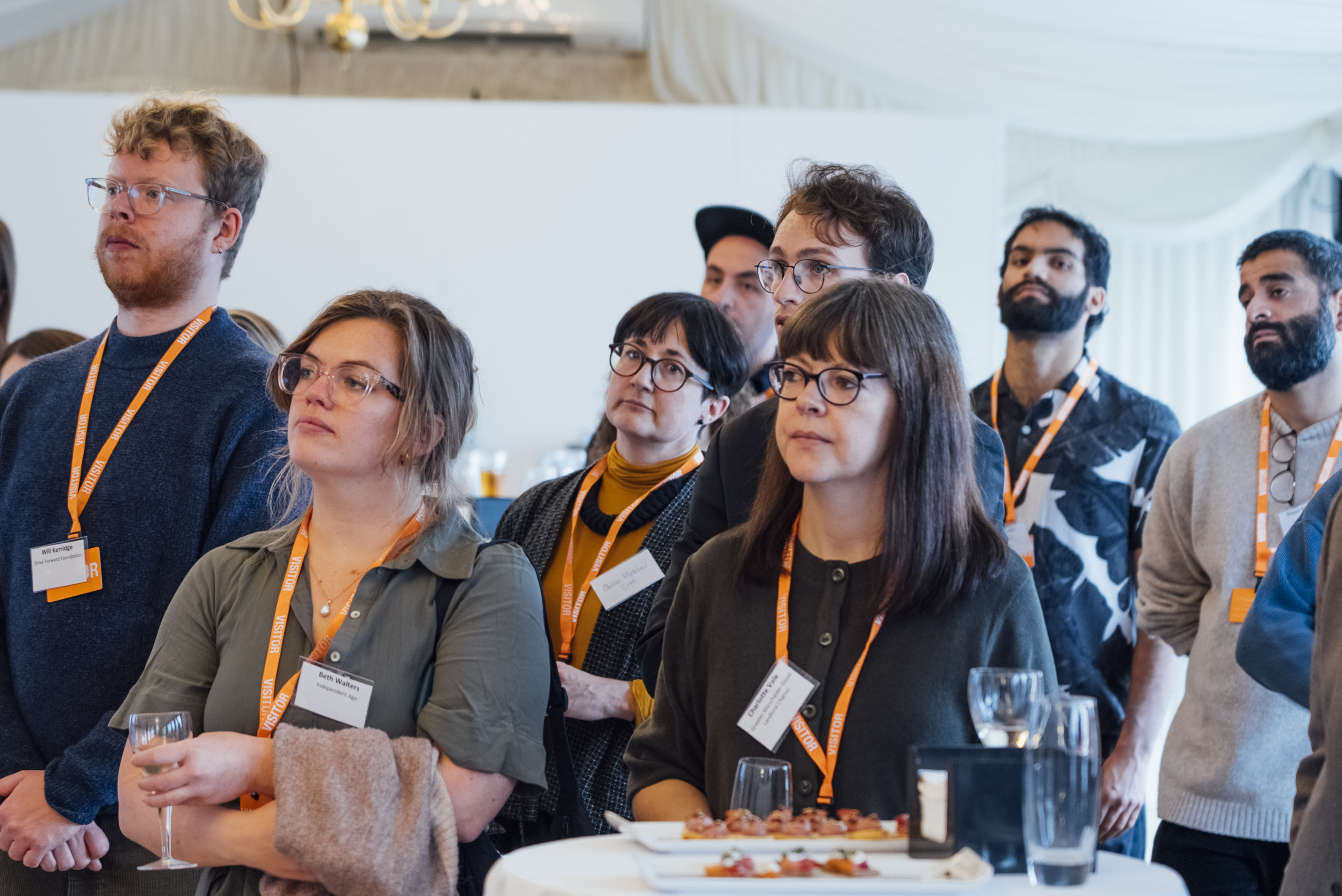 A group of people with lanyards looking off to the side, with drinks and canapes