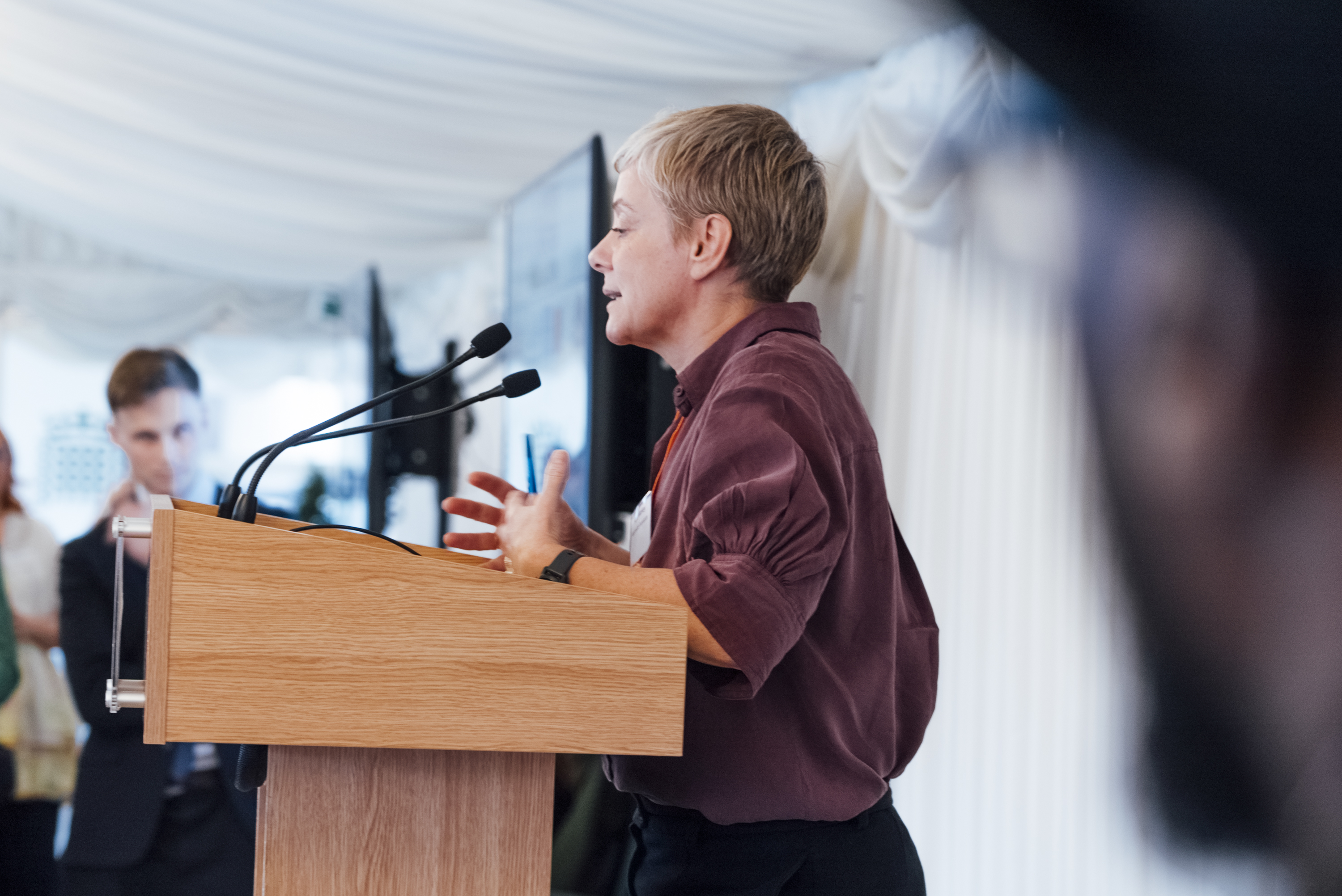 Kate Markey is in focus, speaking at a lectern with a person's face out of focus