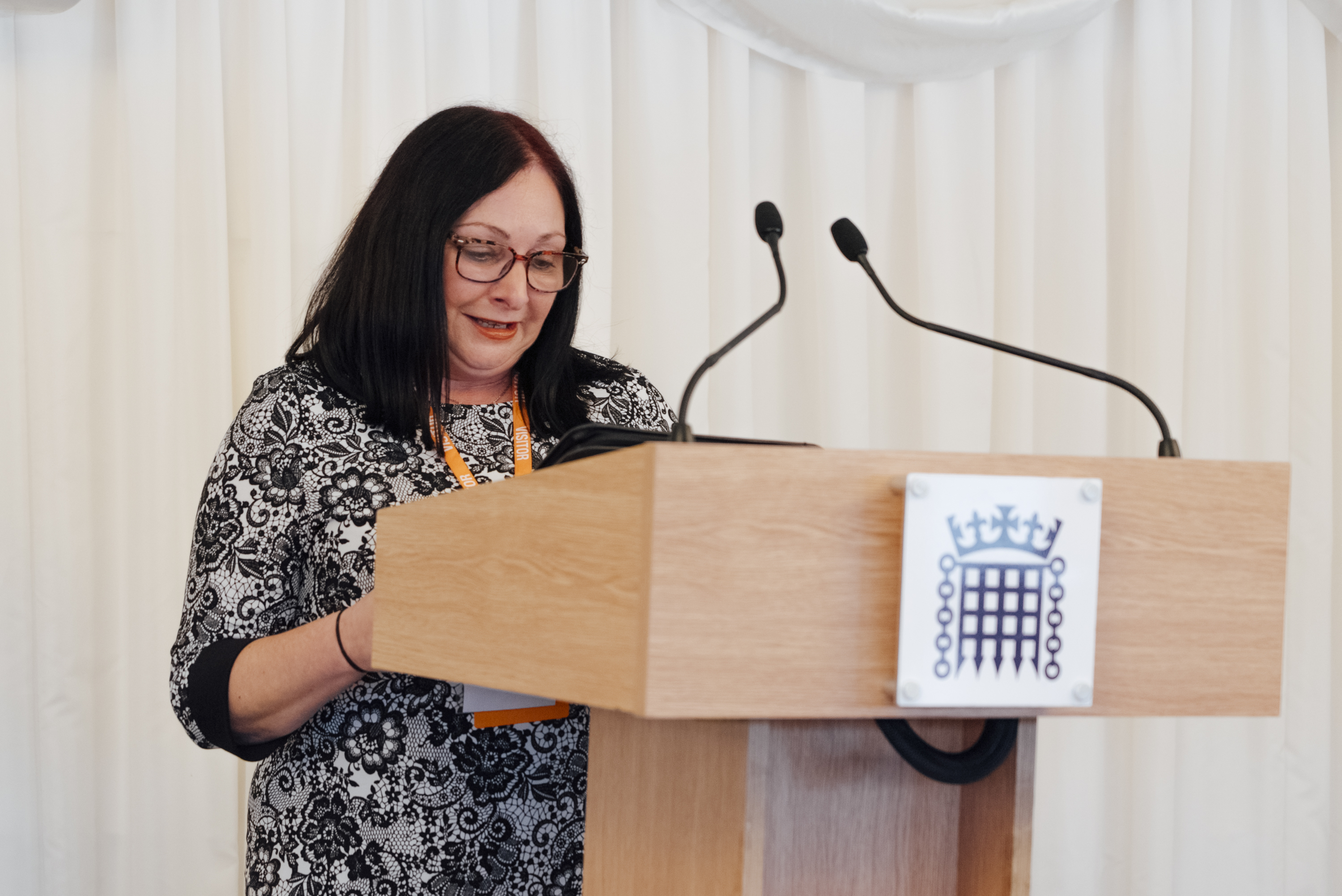 Jen has dark brown hair, an orange lanyard, and a black and white top. She speaks at a podium.