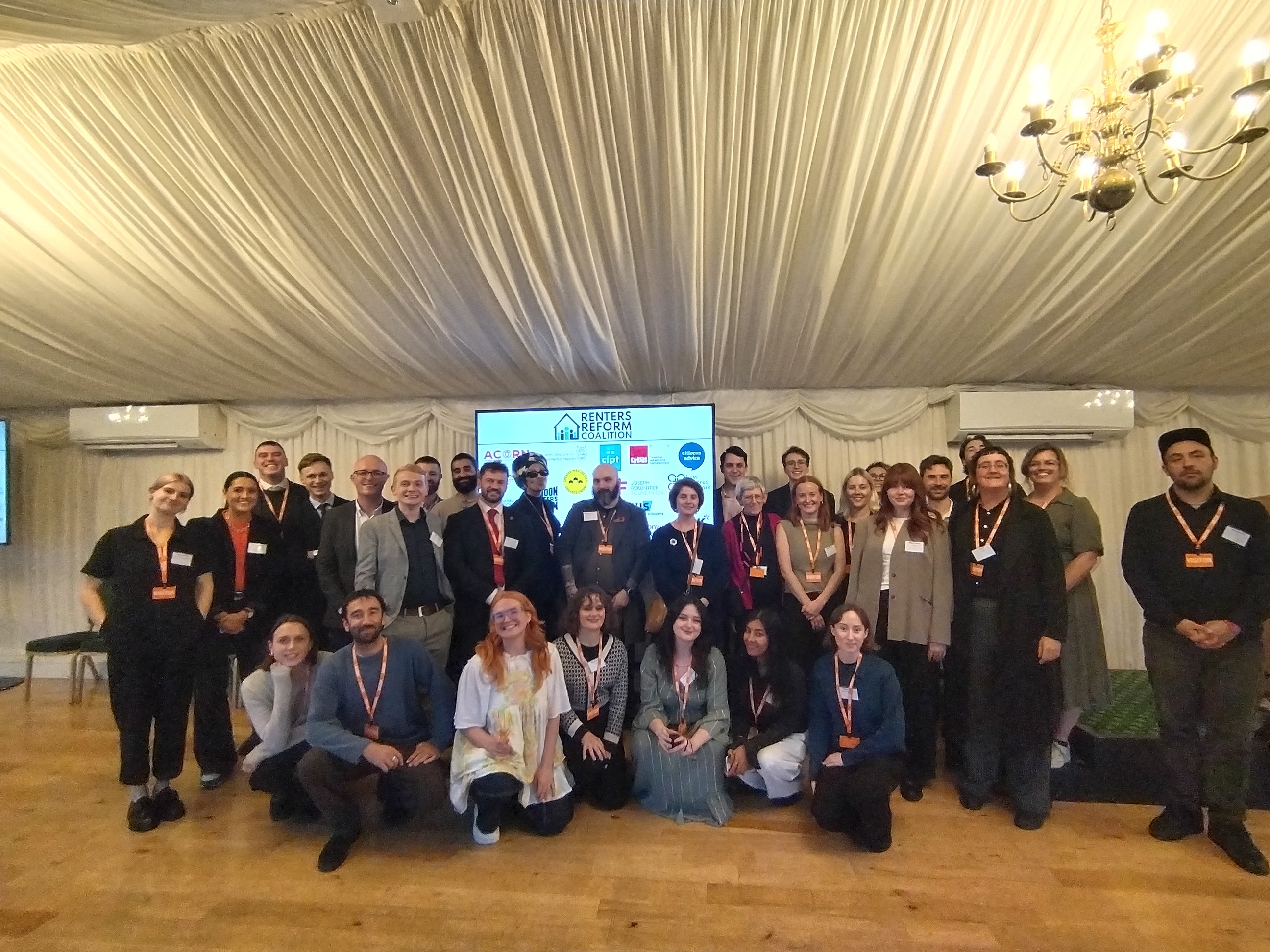 A large group of people smiling at the camera in front of a screen showing the logos of the Renters' Reform Coalition