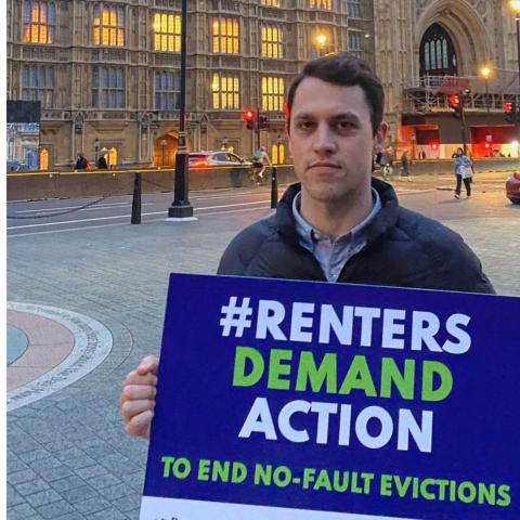 A photo of Paul Shanks, a man with short dark hair holding a placard that reads "#Renters Demand Action to end no-fault evictions"